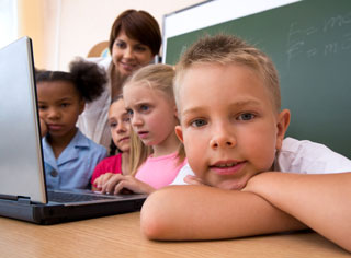 A child looks at the camera while his classmates study a laptop computer