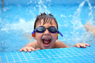 A young boy with swimming goggles grins over the edge of a pool